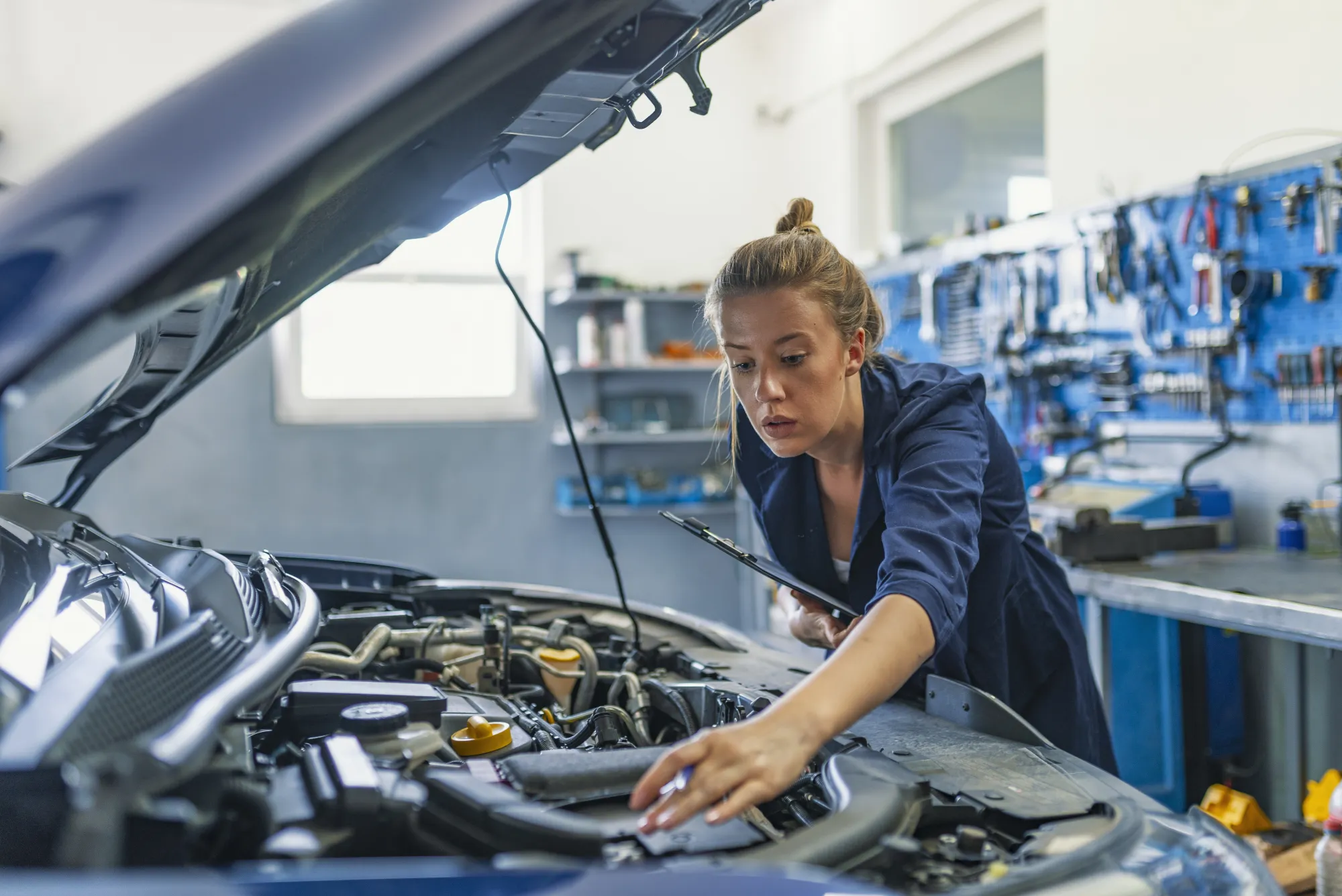 A female mechanic in navy coveralls holds a clipboard while inspecting the engine under a car's open hood in an auto repair shop.