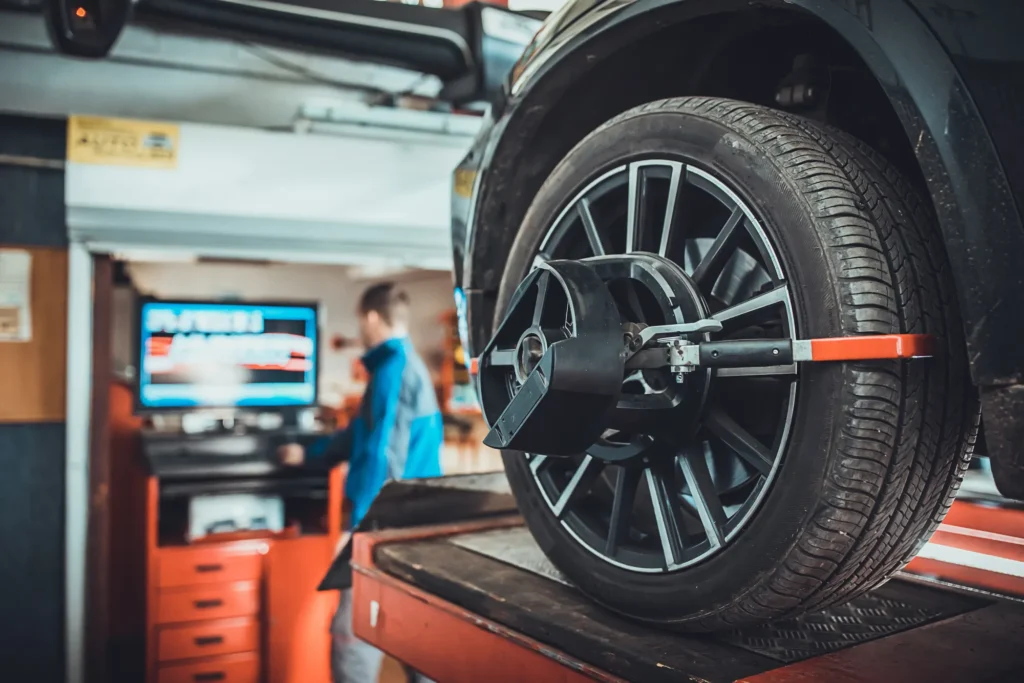 A wheel alignment sensor clamped to the alloy wheel of a car raised on a shop lift, with a technician at the alignment machine in the background.