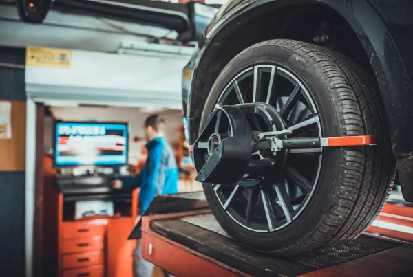 A wheel alignment sensor clamped to the alloy wheel of a car raised on a shop lift, with a technician at the alignment machine in the background.