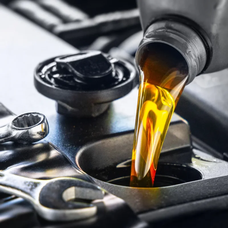 Amber motor oil pouring from a bottle into a car engine's oil fill opening beside two chrome wrenches.