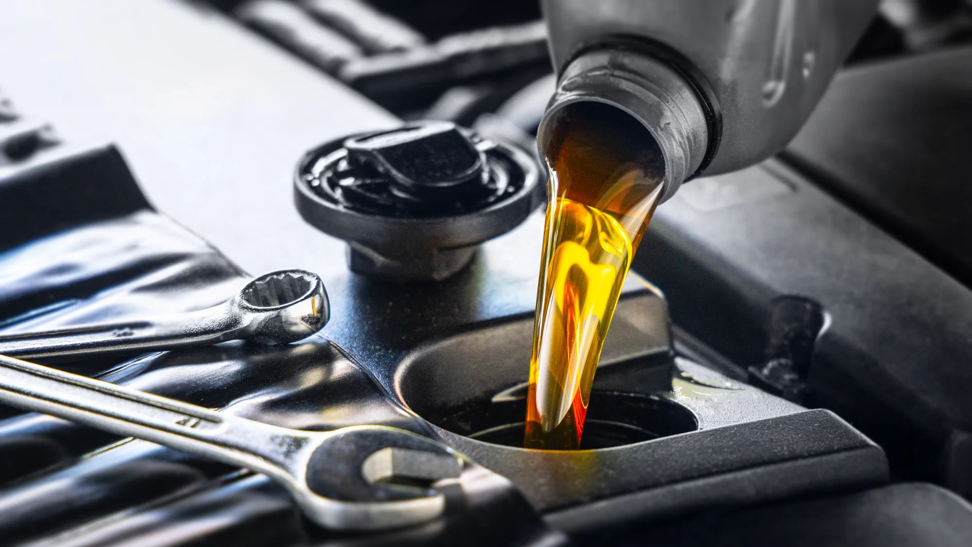 Amber motor oil pouring from a bottle into a car engine's oil fill opening beside two chrome wrenches.