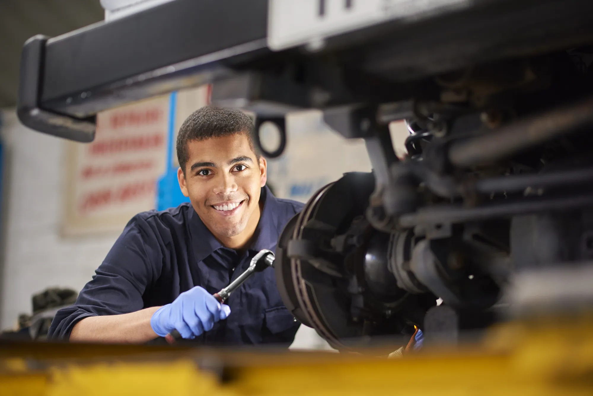 A smiling mechanic in blue gloves holds a socket wrench while working under a car raised on a shop lift, next to the exposed brake assembly.