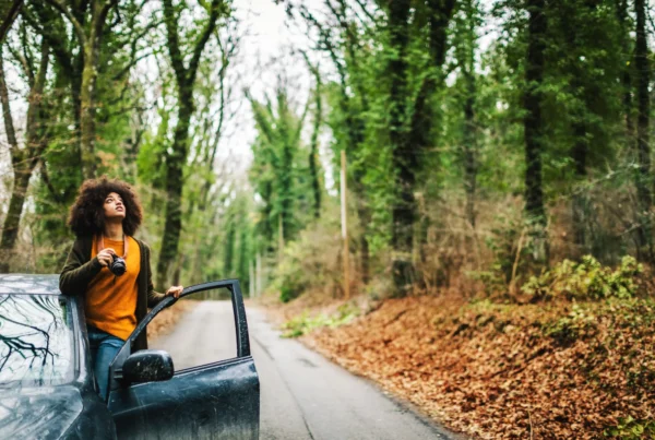 A woman holding a camera stands in the open door of a dark SUV parked on a narrow tree-lined road covered in fallen leaves.