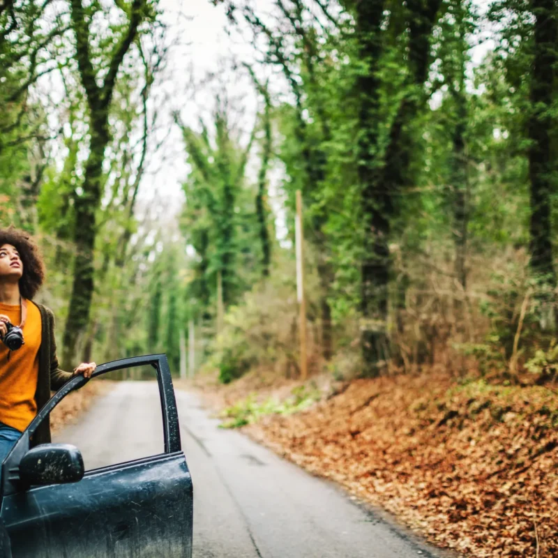 A woman holding a camera stands in the open door of a dark SUV parked on a narrow tree-lined road covered in fallen leaves.