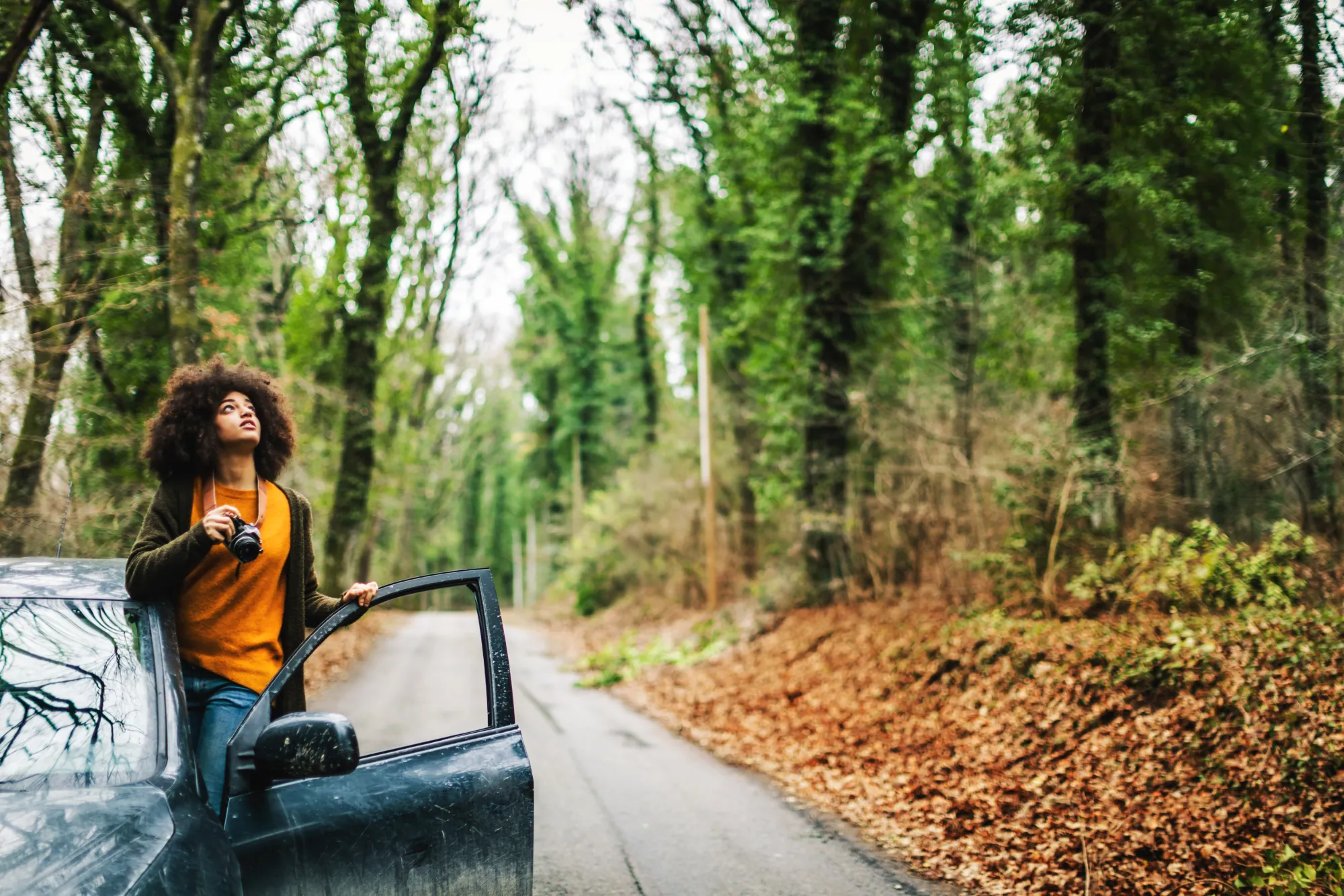 A woman holding a camera stands in the open door of a dark SUV parked on a narrow tree-lined road covered in fallen leaves.