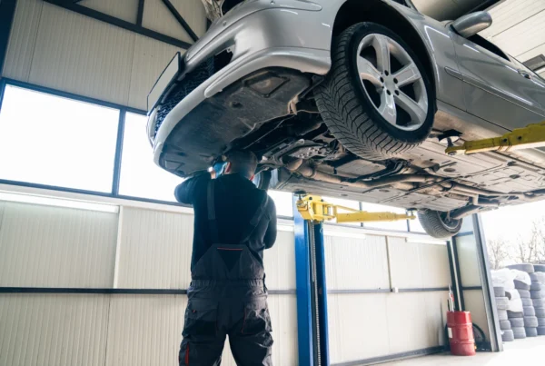 A mechanic in dark coveralls stands beneath a silver car raised on a lift, working on its undercarriage in a repair shop bay.