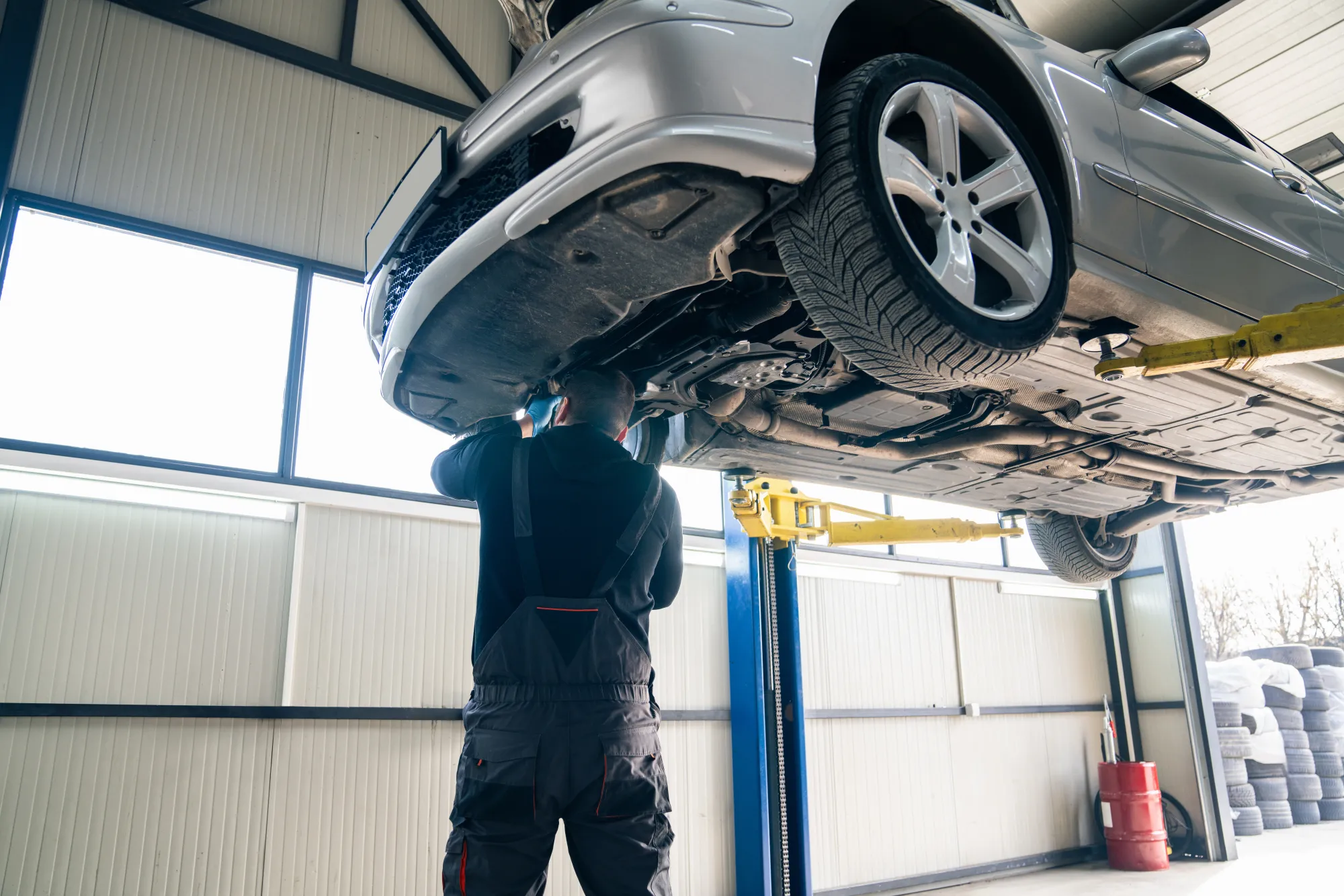 A mechanic in dark coveralls stands beneath a silver car raised on a lift, working on its undercarriage in a repair shop bay.