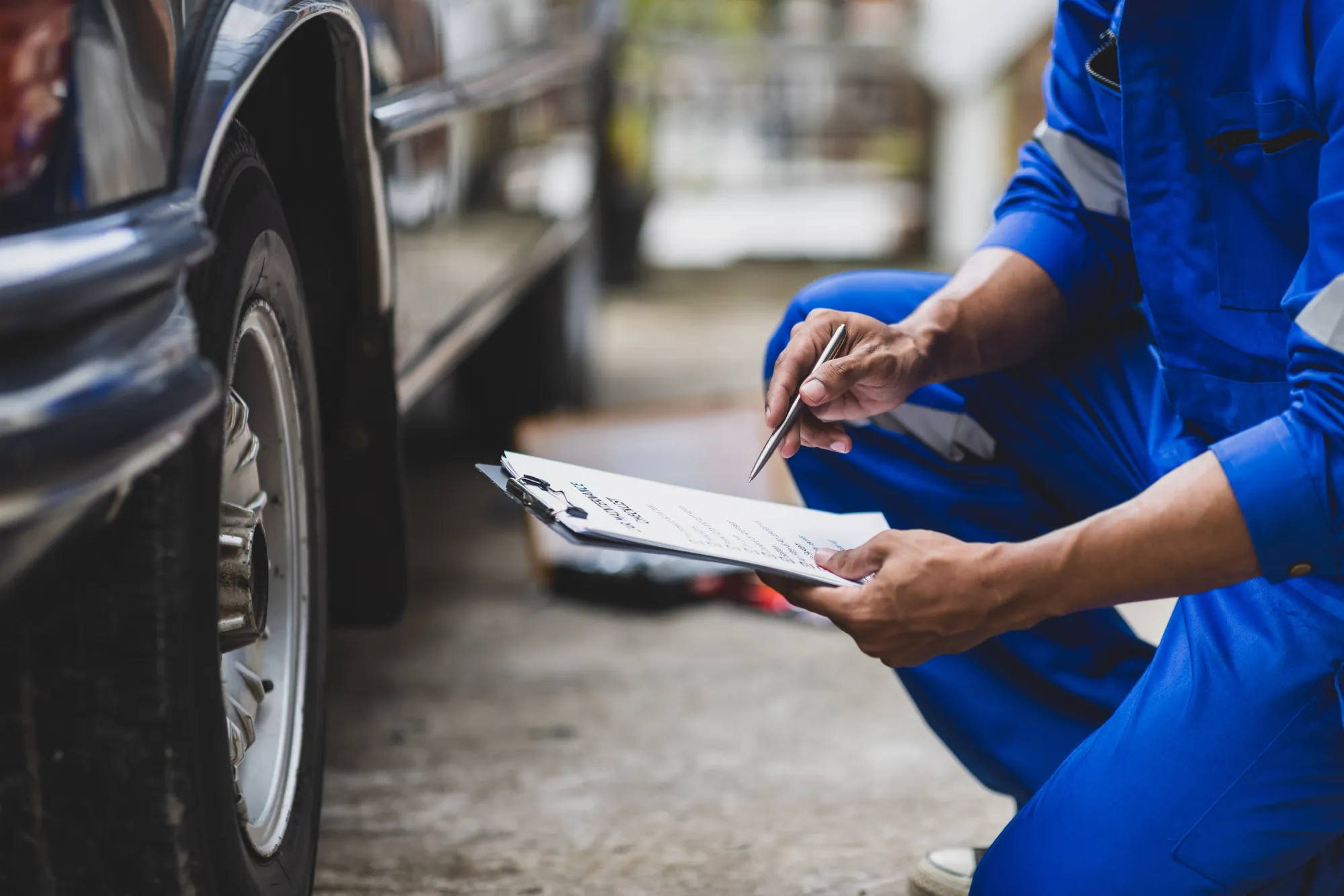 A mechanic in blue coveralls kneels next to a car's front tire, filling out a car maintenance checklist on a clipboard.